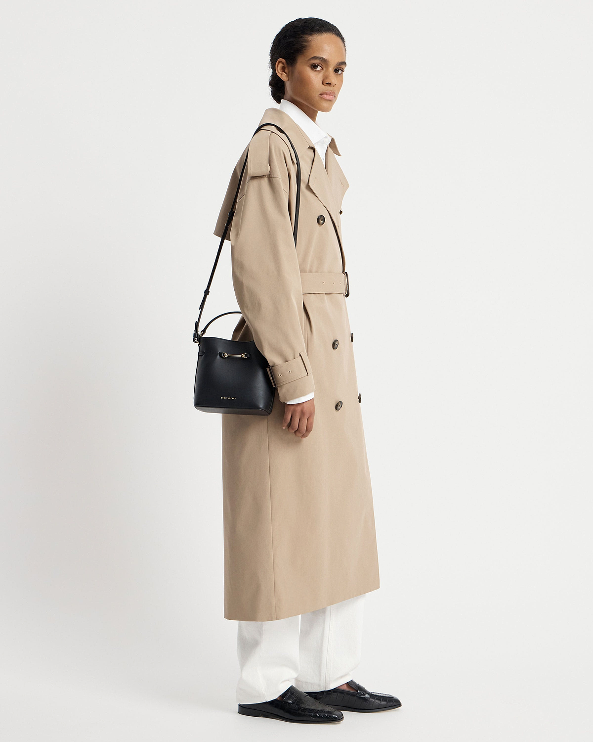 Model in beige trench coat carrying black Strathberry Corda Bucket Mini shoulder bag.