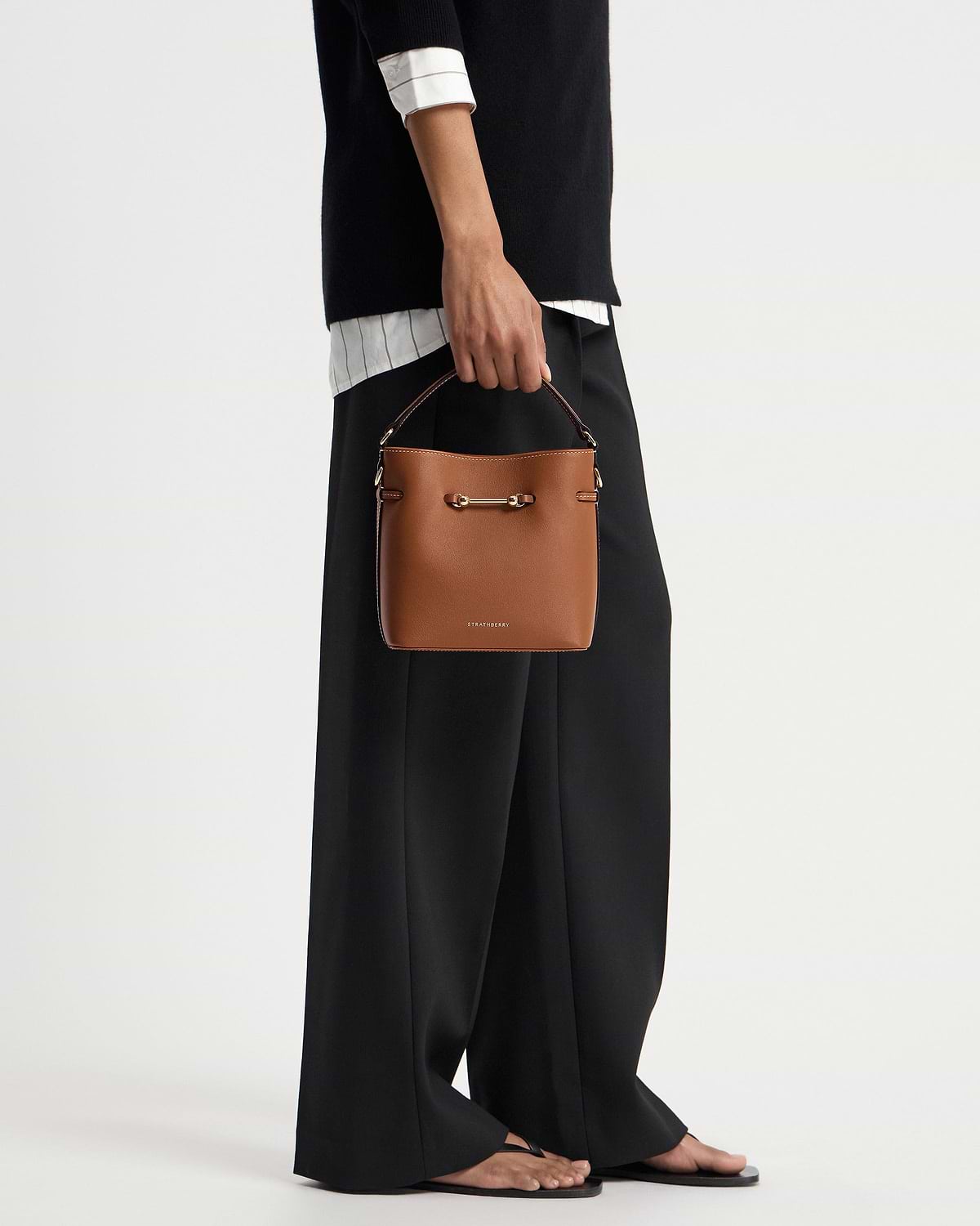 Hand holding chestnut Strathberry bucket bag with gold hardware, model in black trousers.