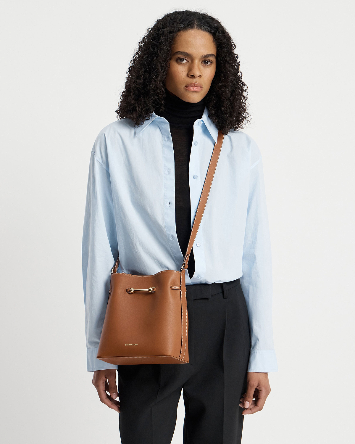 Model wearing chestnut Corda bucket bag by Strathberry, cross-body over blue shirt.