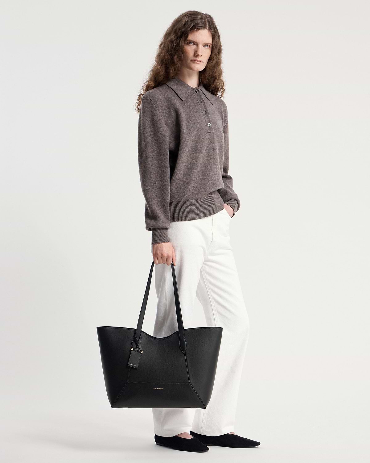 Model holding black Strathberry Mosaic Cabas tote by handles, wearing white trousers.