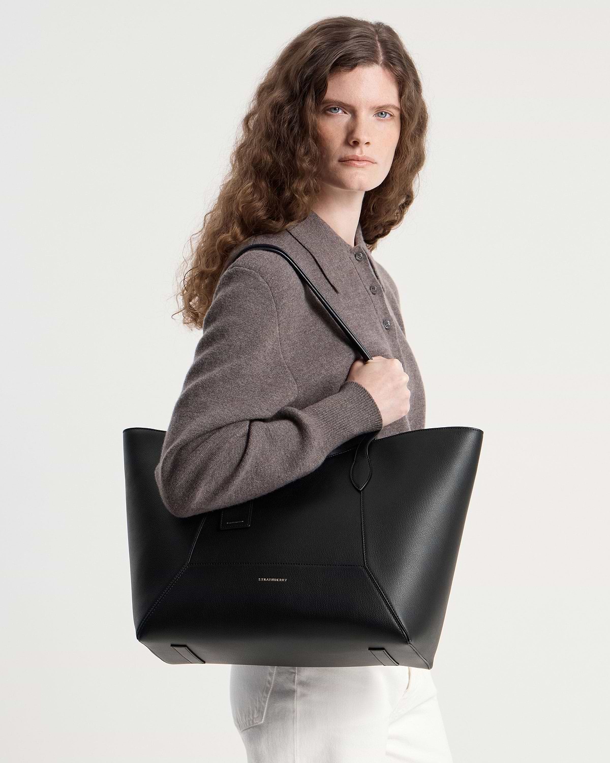 Model wearing brown jumper holding black Strathberry tote bag over shoulder.