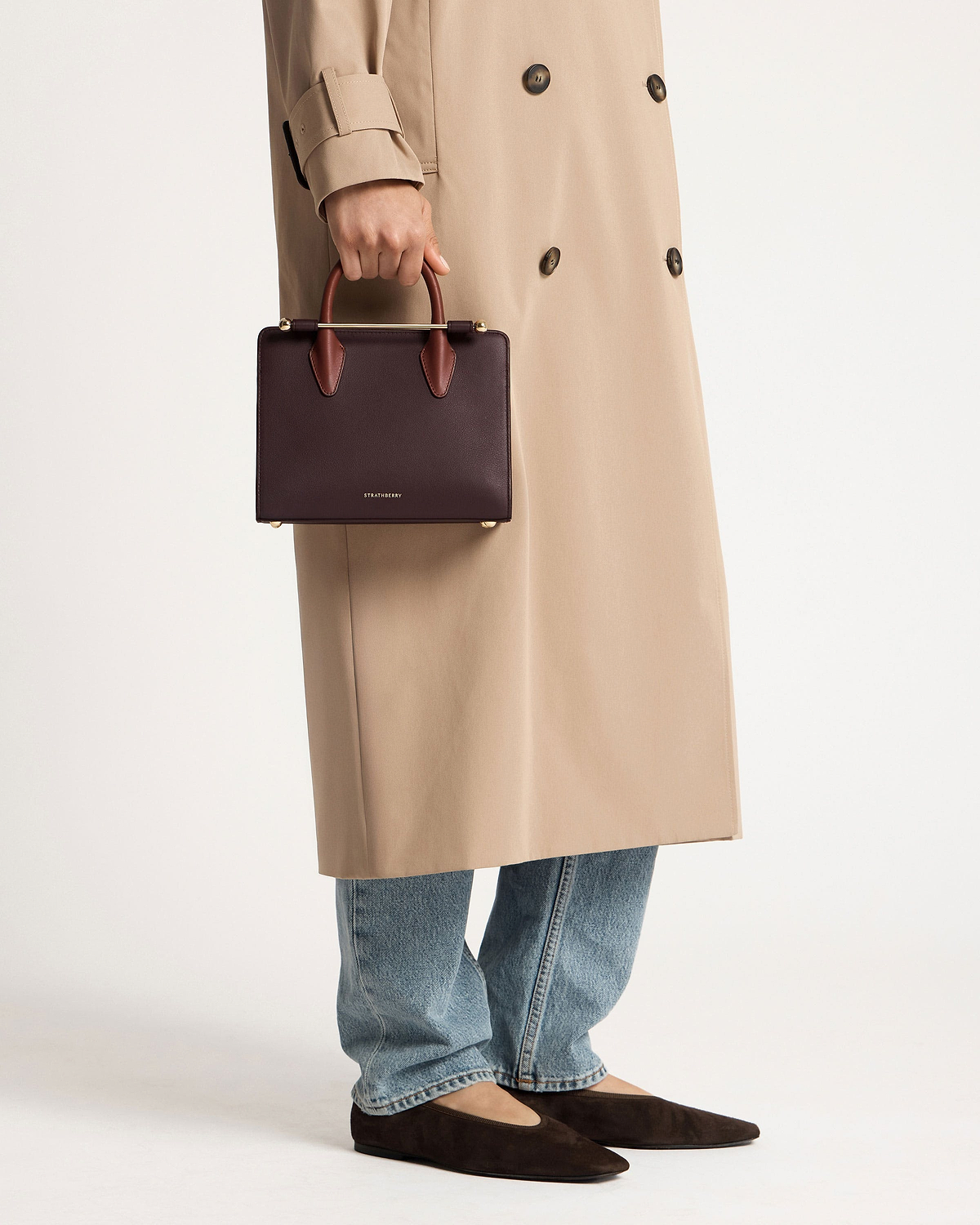 Hand holding Strathberry burgundy mini tote with chestnut handles, model in trench and jeans.