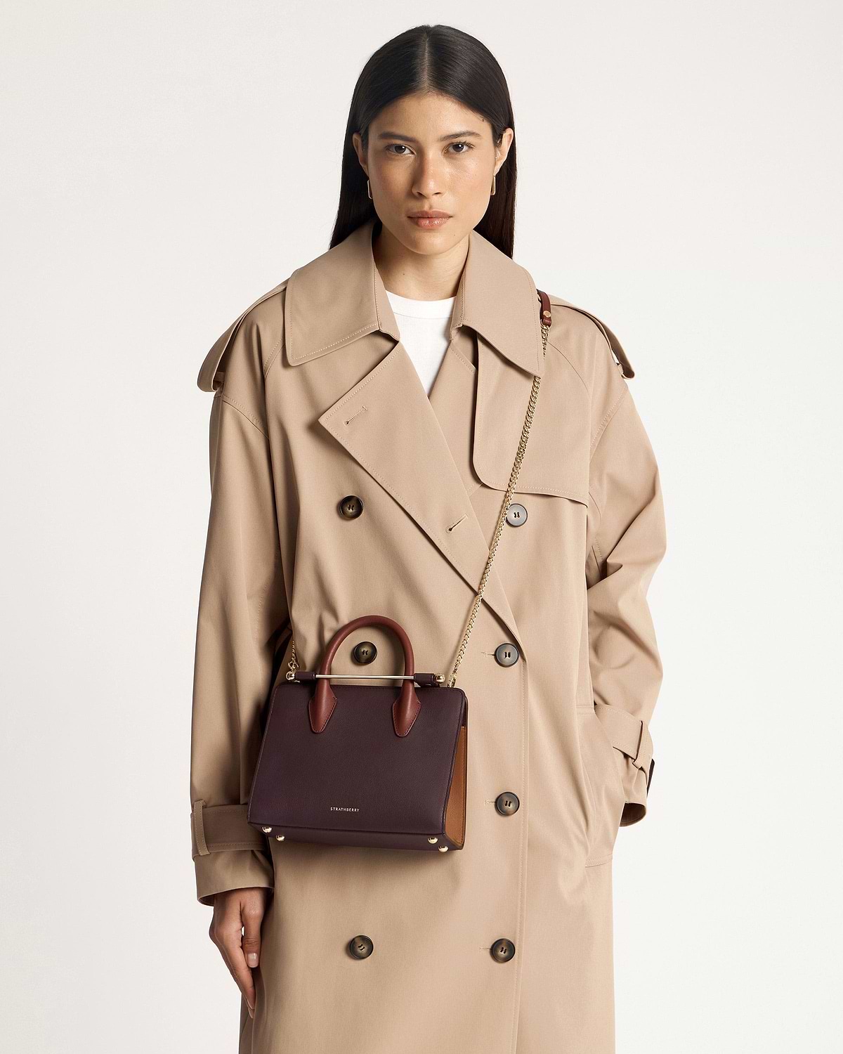 Model wearing beige trench coat with burgundy Strathberry mini tote, chestnut handles, walnut side panel.