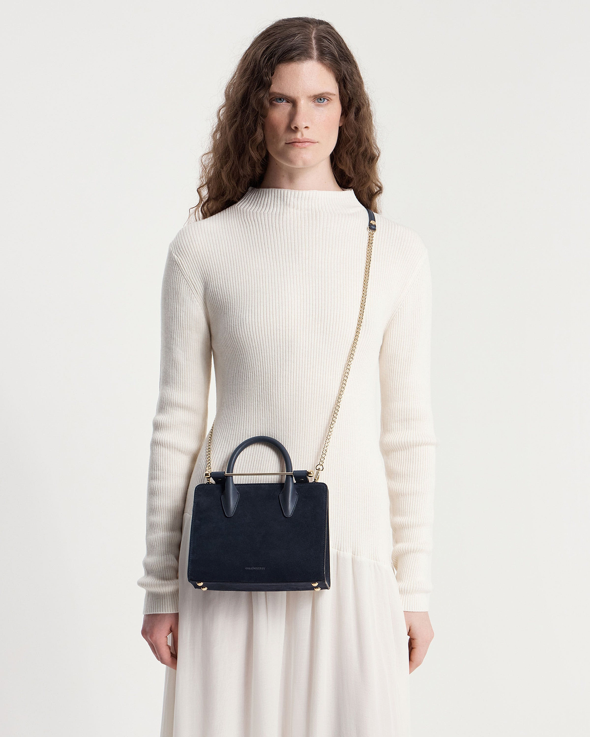 Model wearing cream ribbed top carrying a navy suede mini tote with chain strap.