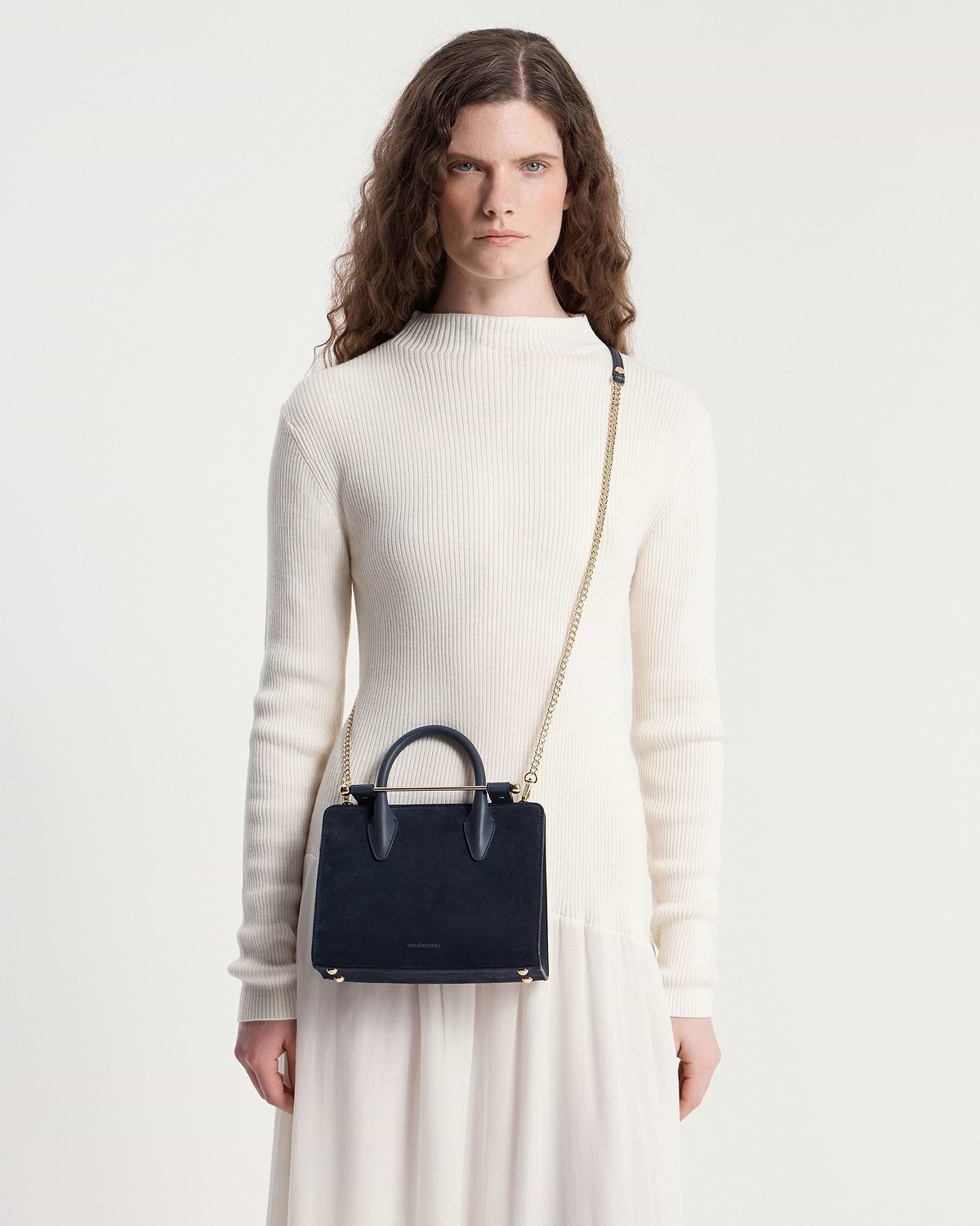 Model wearing cream ribbed top carrying a navy suede mini tote with chain strap.