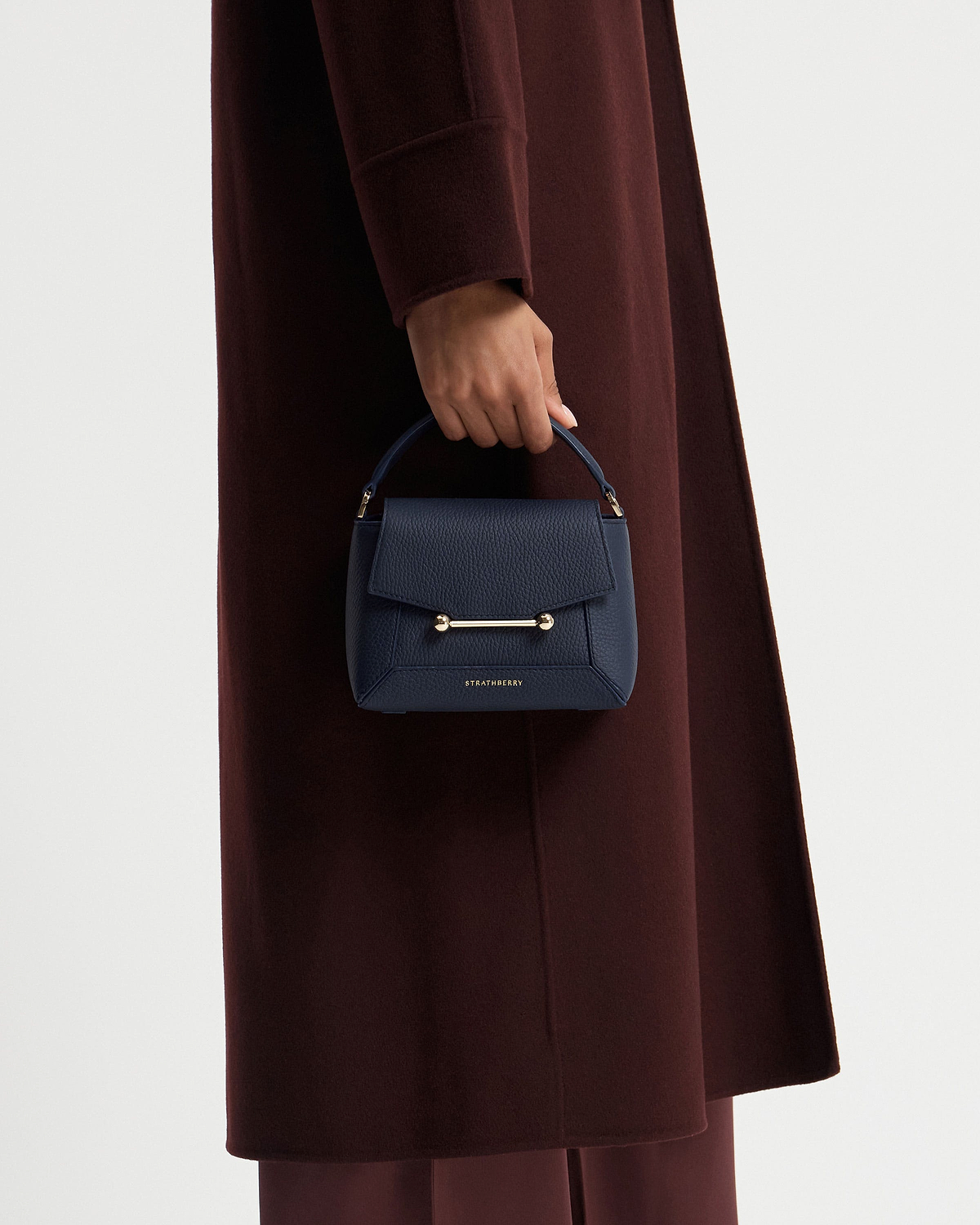 Model's hand holding navy Strathberry Mosaic Nano bag against coat.