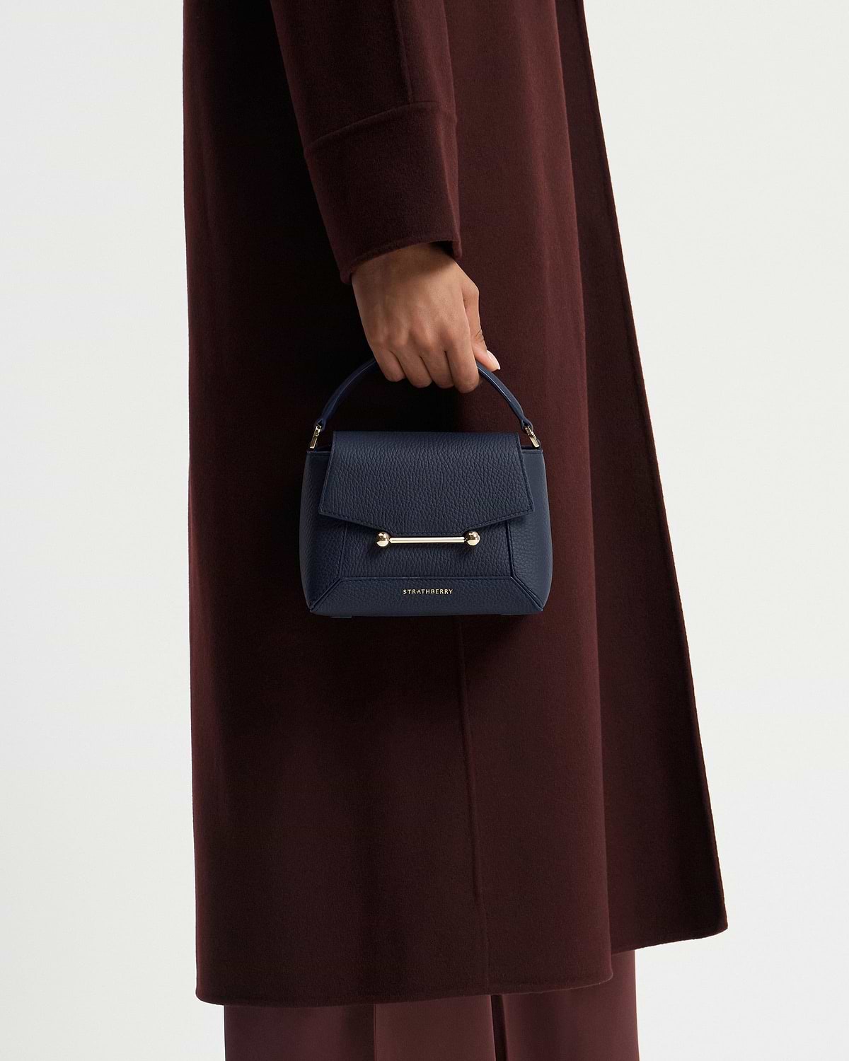 Model's hand holding navy Strathberry Mosaic Nano bag against coat.
