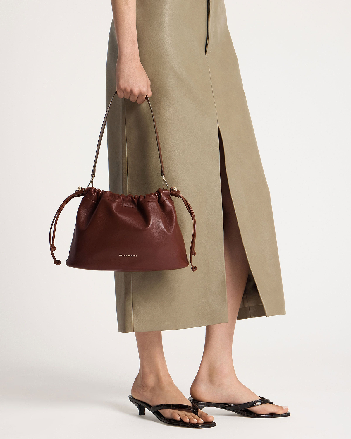 Model holding a Strathberry Charlotte midi drawstring bag in walnut, lower legs visible.
