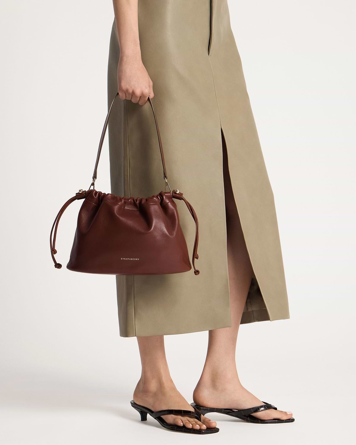 Model holding a Strathberry Charlotte midi drawstring bag in walnut, lower legs visible.