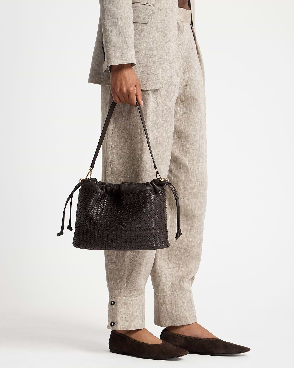 Model holding chocolate woven leather drawstring bag with beige suit trousers and flats