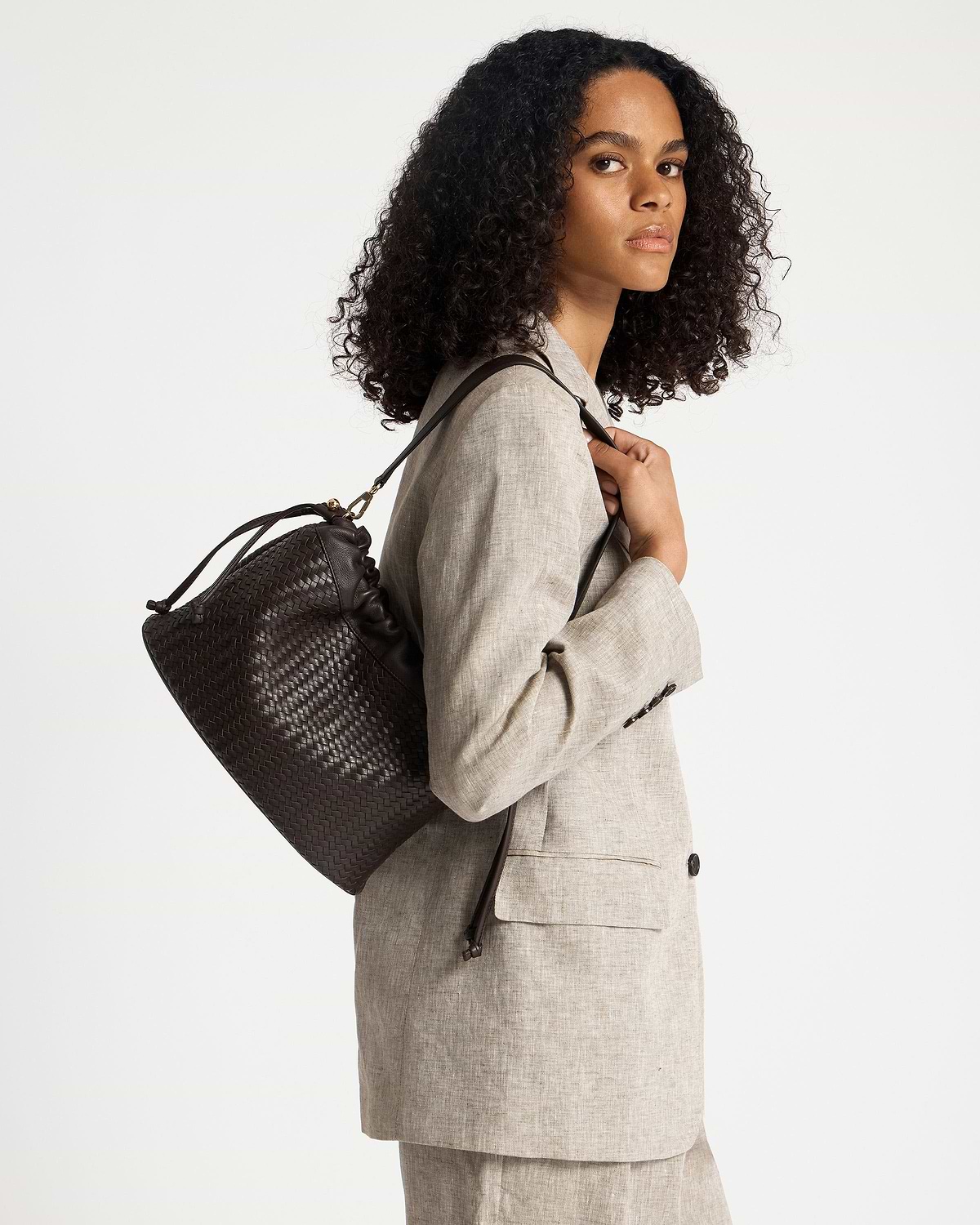 Woman in beige blazer carrying chocolate woven leather drawstring bag over shoulder.