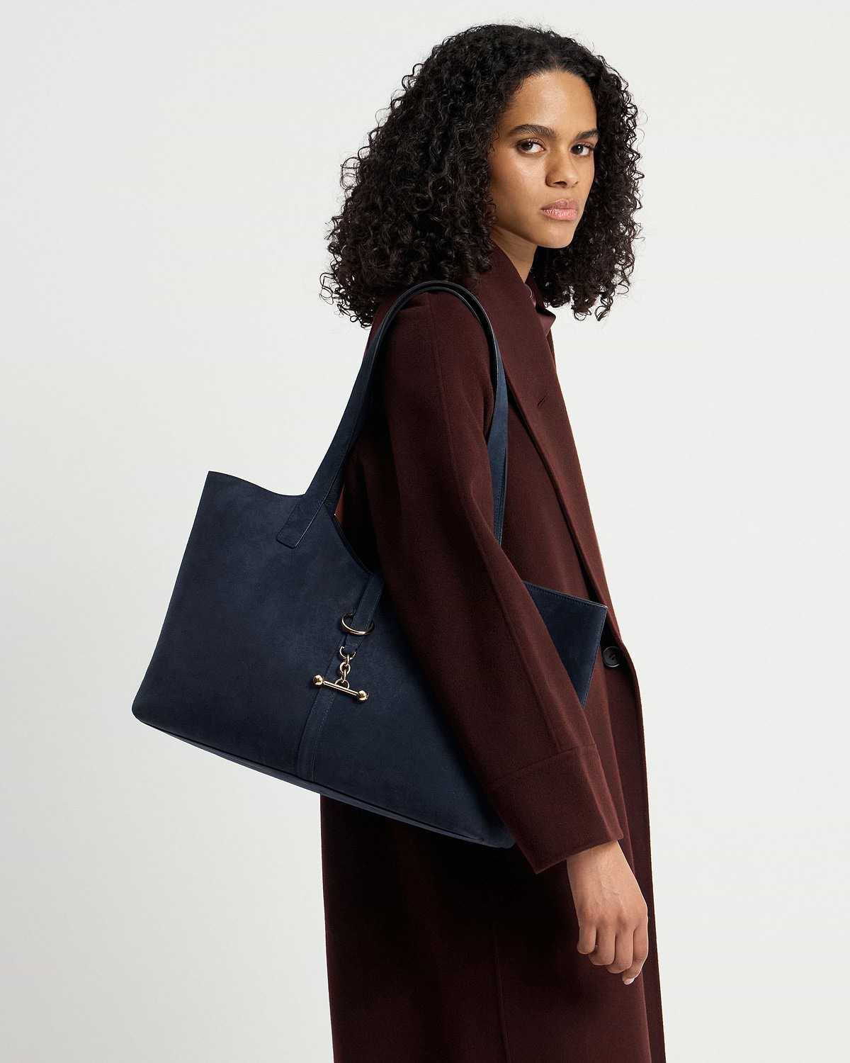 Model wearing burgundy coat carrying navy suede Kite tote over shoulder.