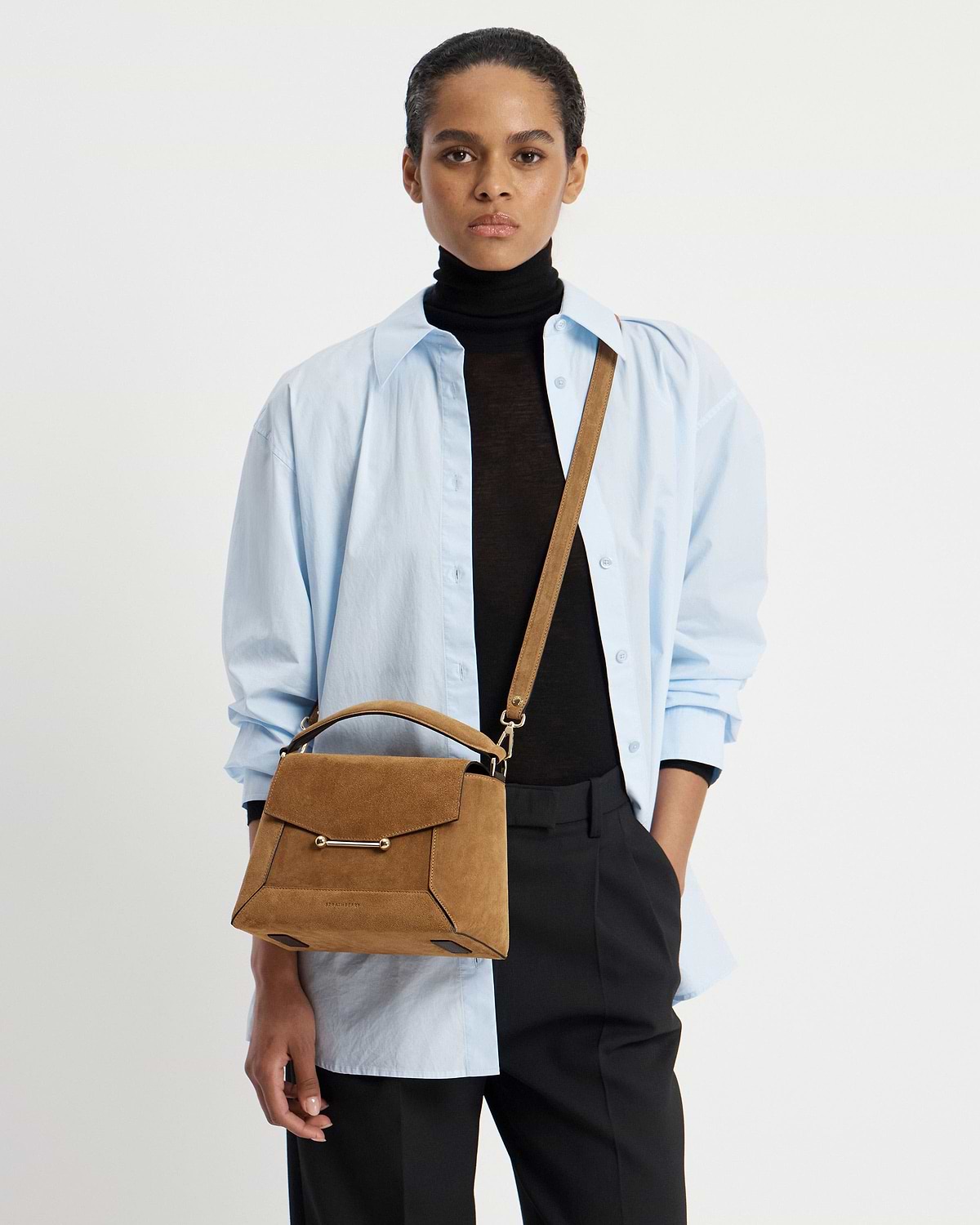 Model wearing light blue shirt and black roll-neck carrying toffee suede mosaic bag.