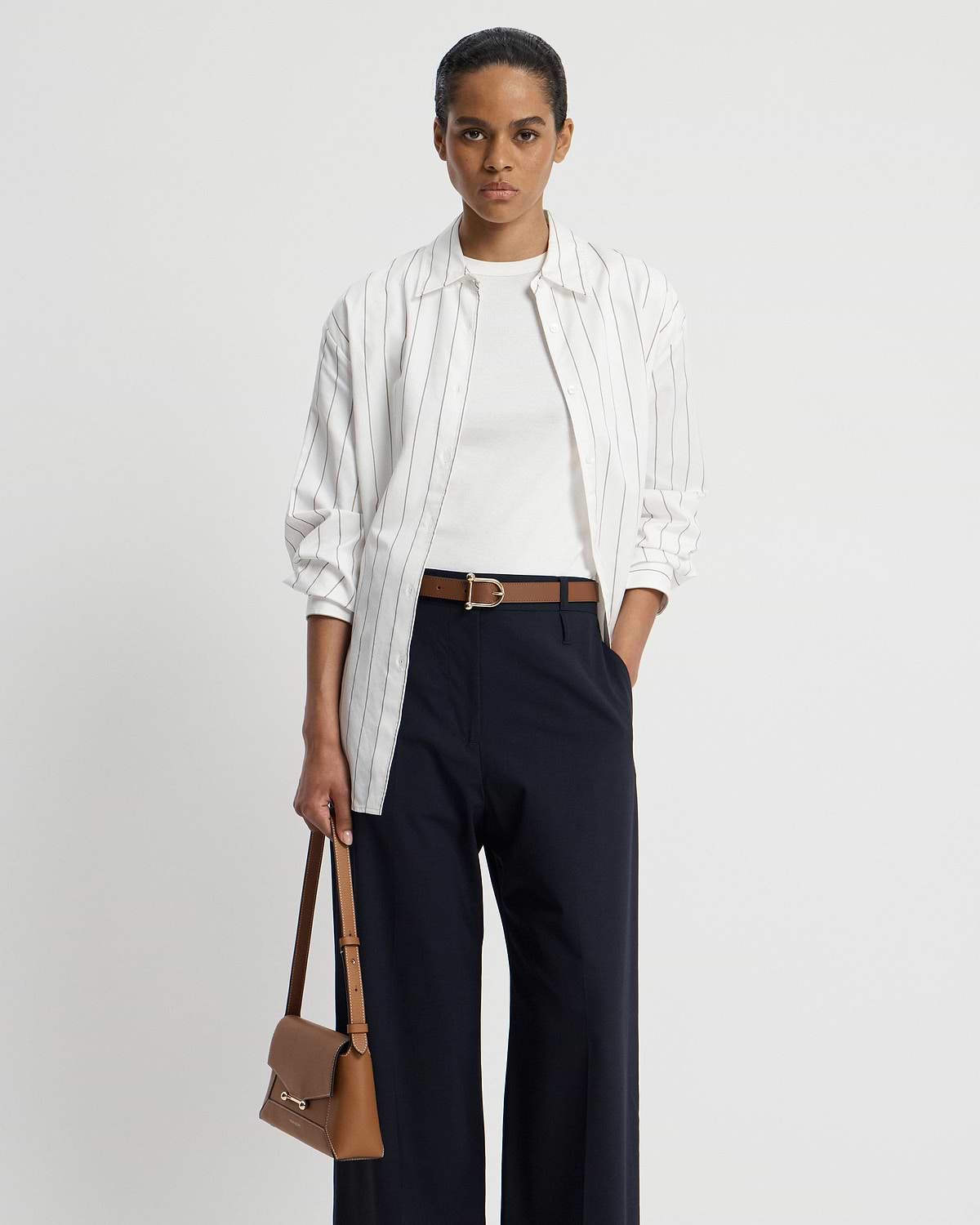 Model wearing hazelnut leather belt with gold buckle over navy trousers.