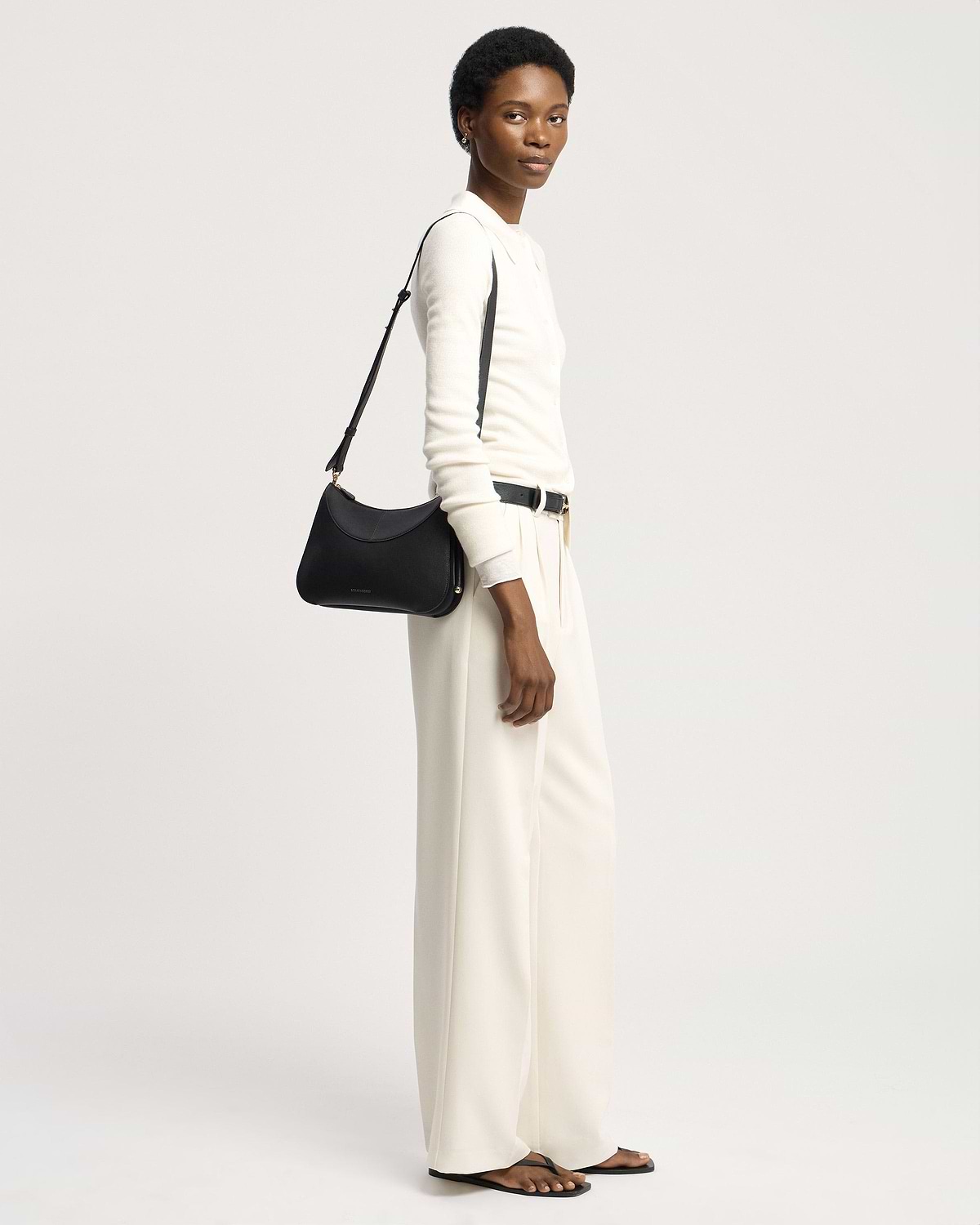 Model in white outfit carrying black Strathberry Nest shoulder bag on shoulder.