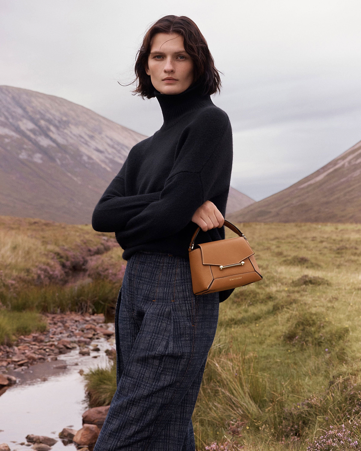 Woman in black jumper holding tan Strathberry Mosaic Nano bag with vanilla stitching.