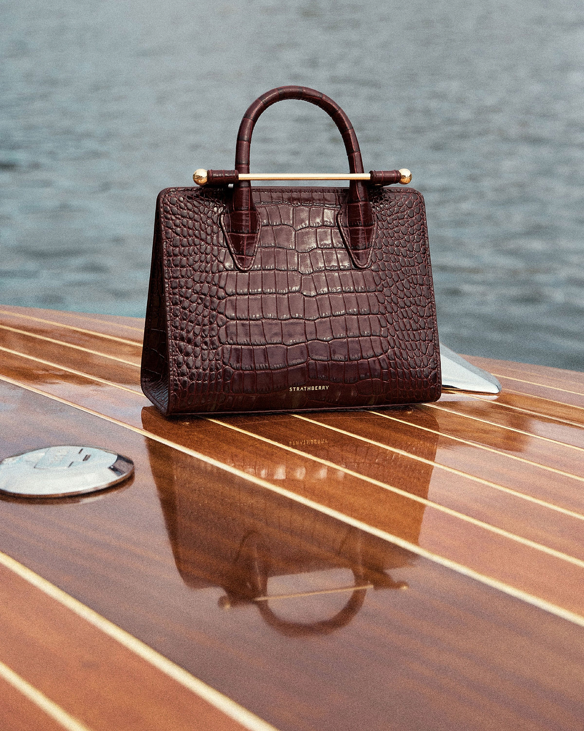 Strathberry croc-embossed mini tote resting on a wooden boat deck