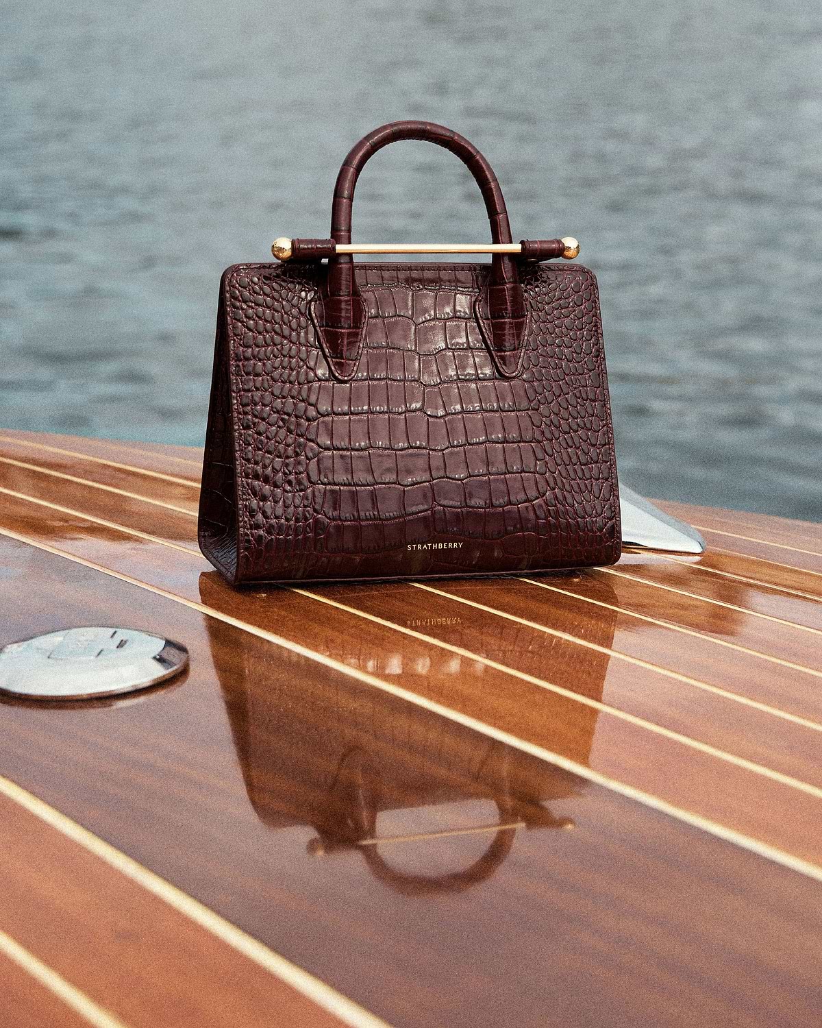 Strathberry croc-embossed mini tote resting on a wooden boat deck