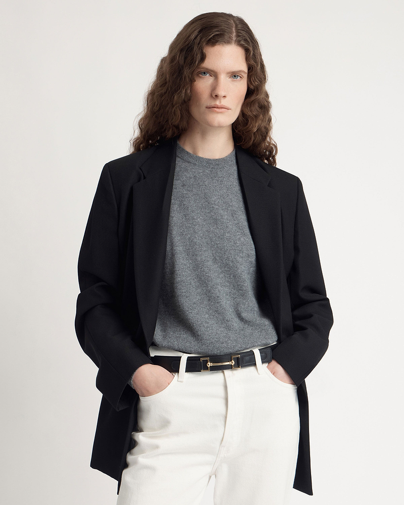 Model wearing black leather belt with gold hardware over white trousers and grey sweater.