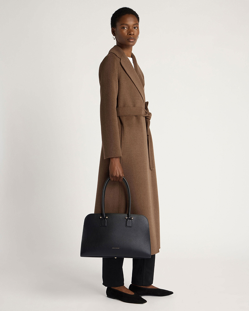 Woman holding black Georgia Maxi tote bag while wearing a brown belted coat.