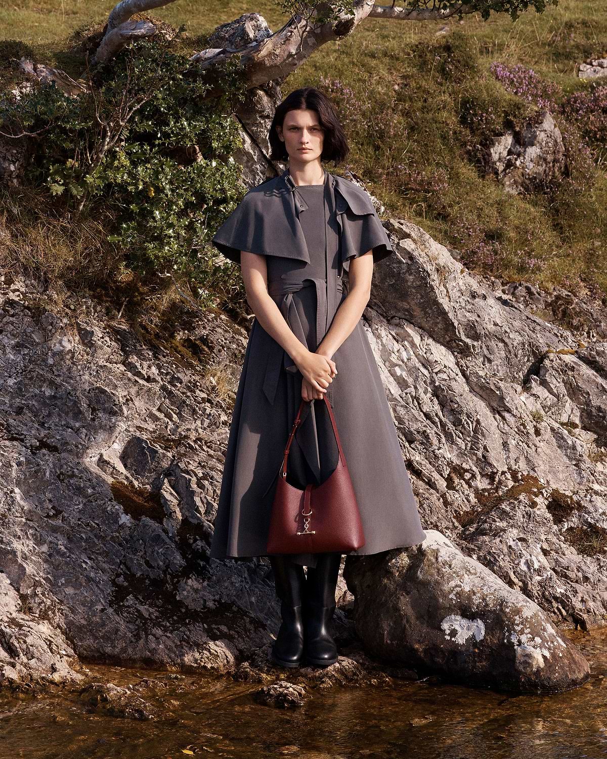 Woman standing on rocky shore holding an oxblood Kite Hobo shoulder bag.