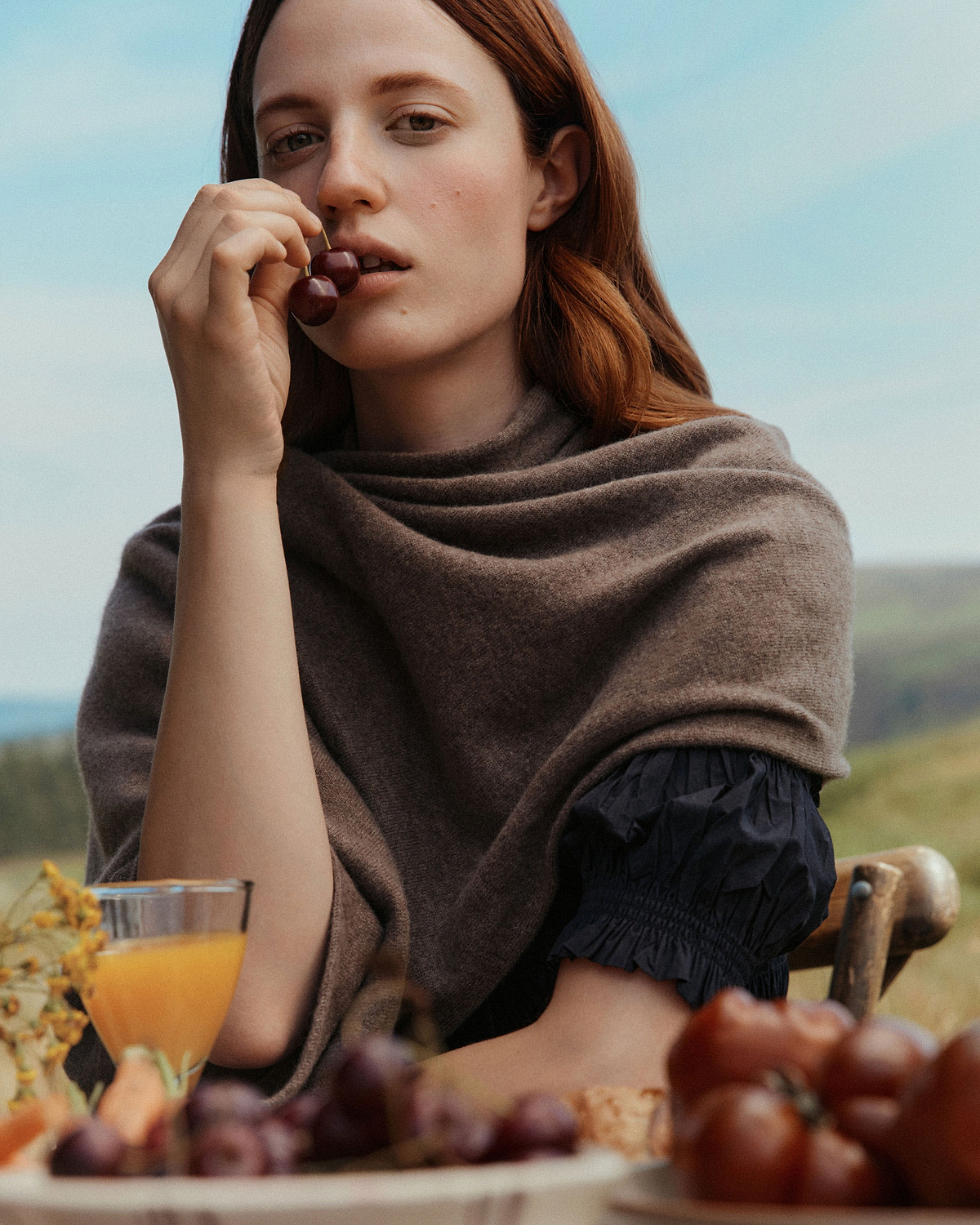 Woman wearing otter-coloured cashmere travel wrap draped over shoulders, holding cherries.