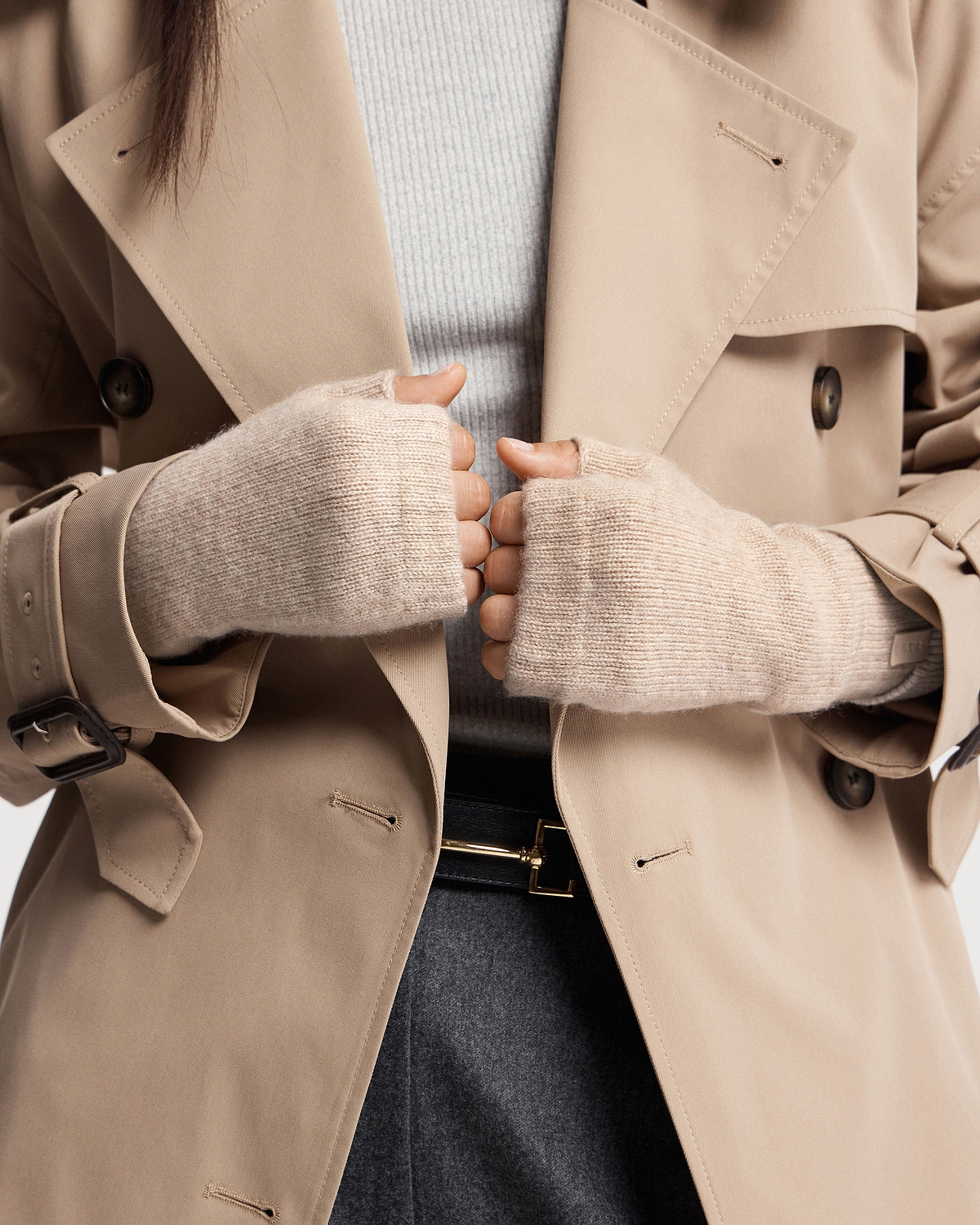 Hands in oat fingerless cashmere gloves holding beige trench coat lapels.