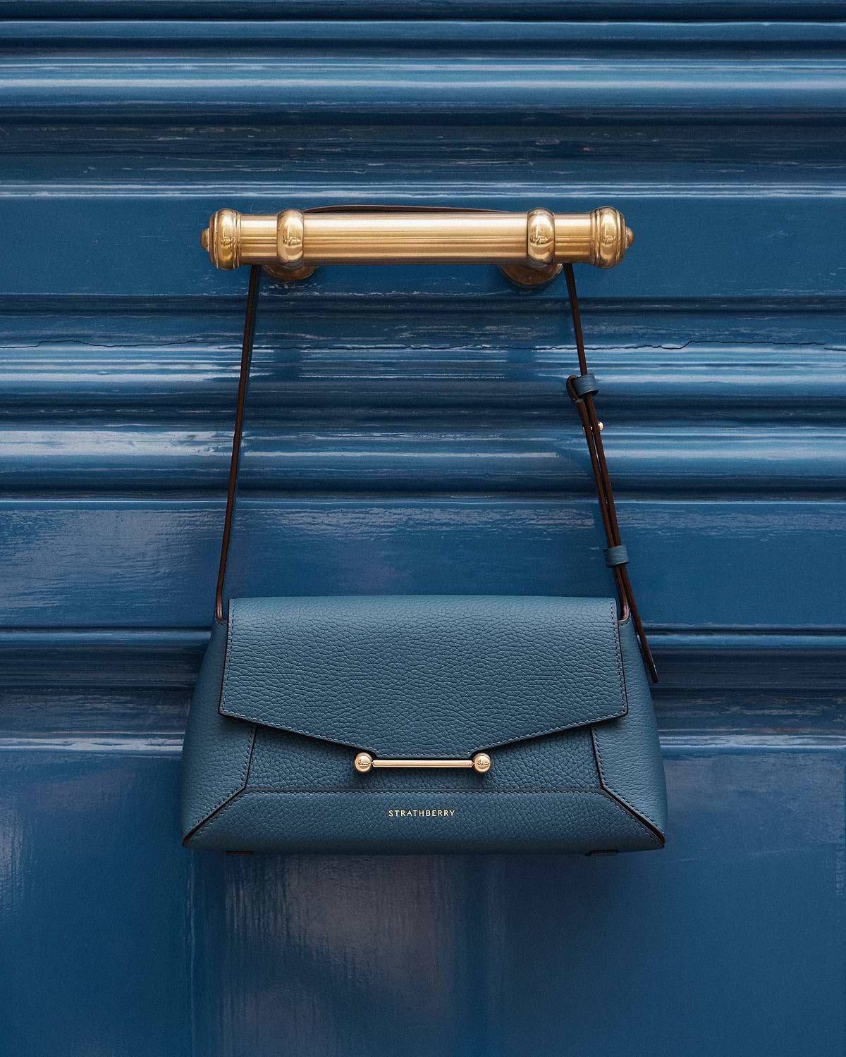 Strathberry Mosaic shoulder bag in loch blue hanging from brass door handle.