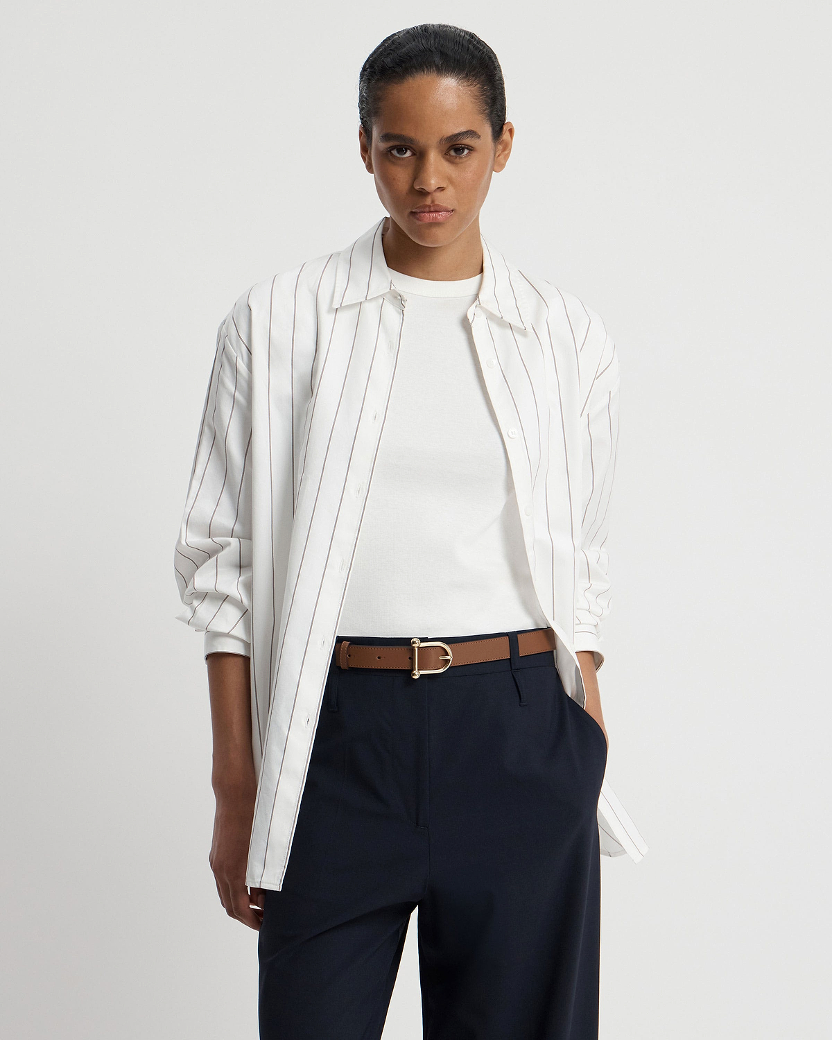 Model wearing white striped shirt, white T-shirt, navy trousers and hazelnut belt with gold buckle.