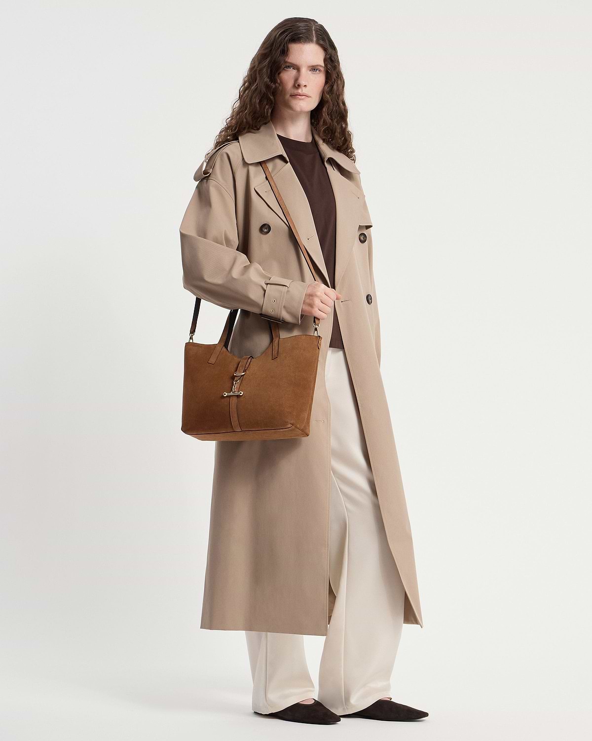 Model in a beige trench coat carrying a tan suede tote bag at their side.