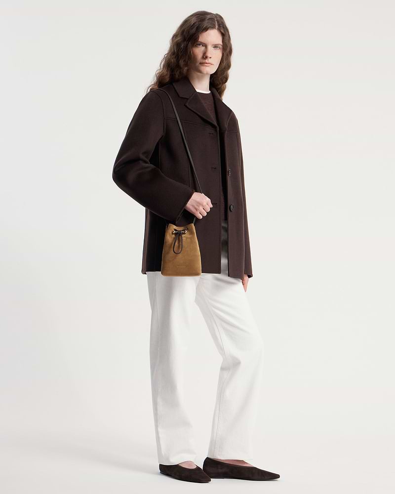 Model wearing dark brown coat holding a toffee suede drawstring pouch with dark shoulder strap.