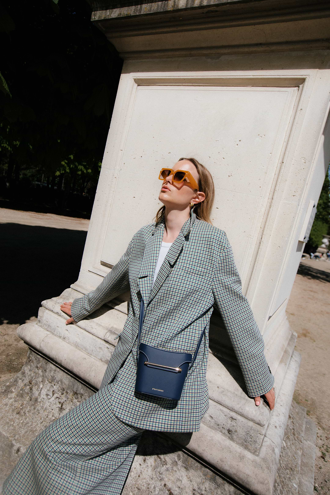 A woman in a suit and sunglasses leaning against a wall