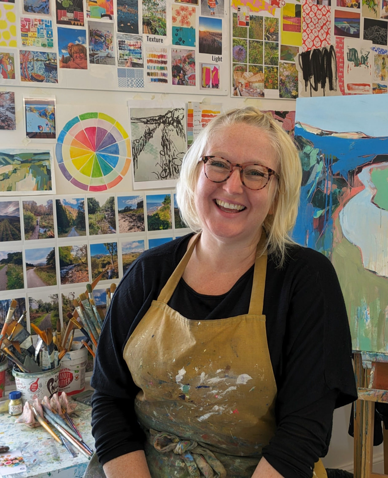 A woman in an apron is smiling in front of a painting
