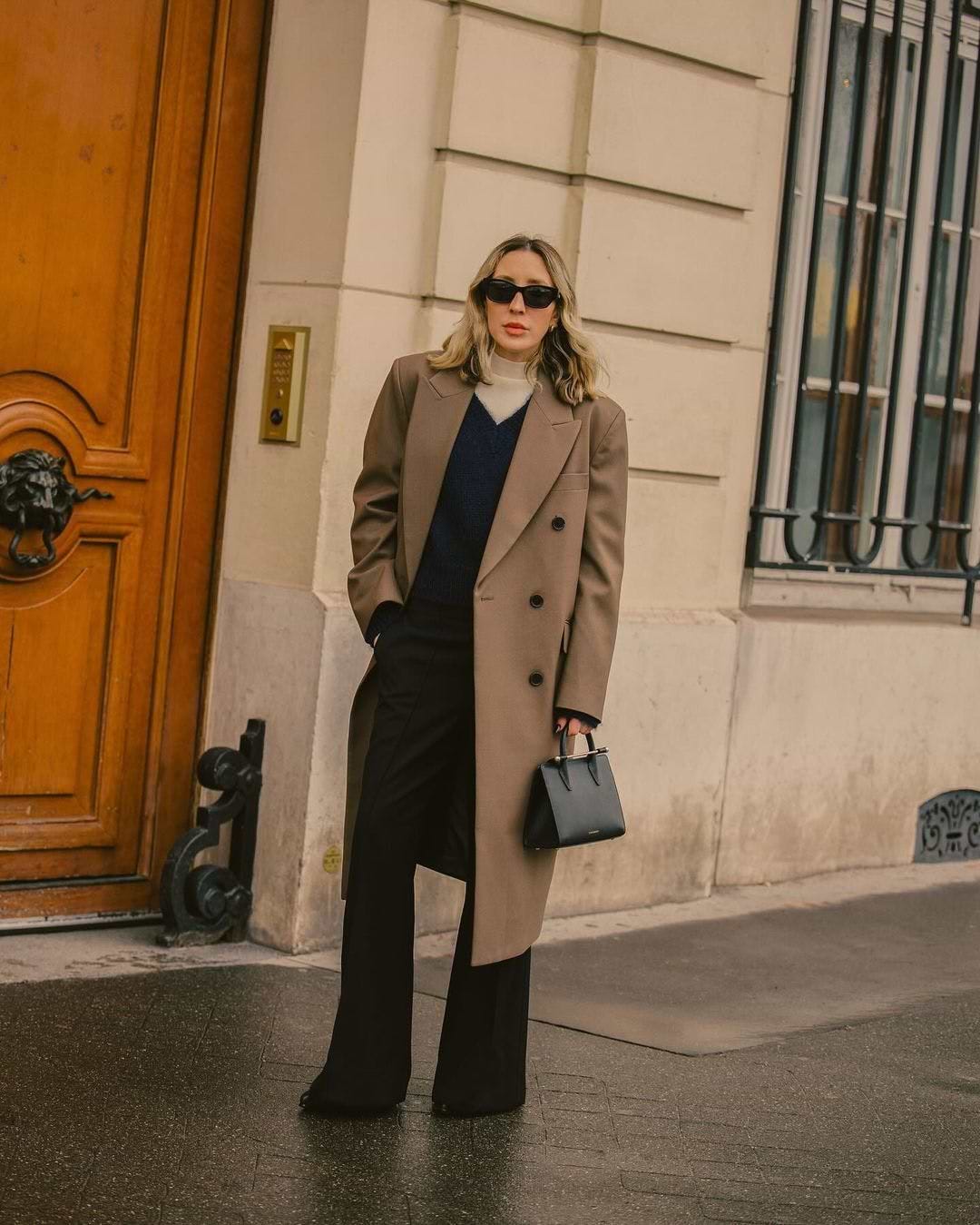 A woman in a tan coat and black pants stands in front of a door