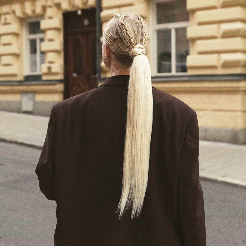 Person with long blonde hair extensions in ponytail and braid, wearing a dark blazer, stands on a street with beige buildings in the background.