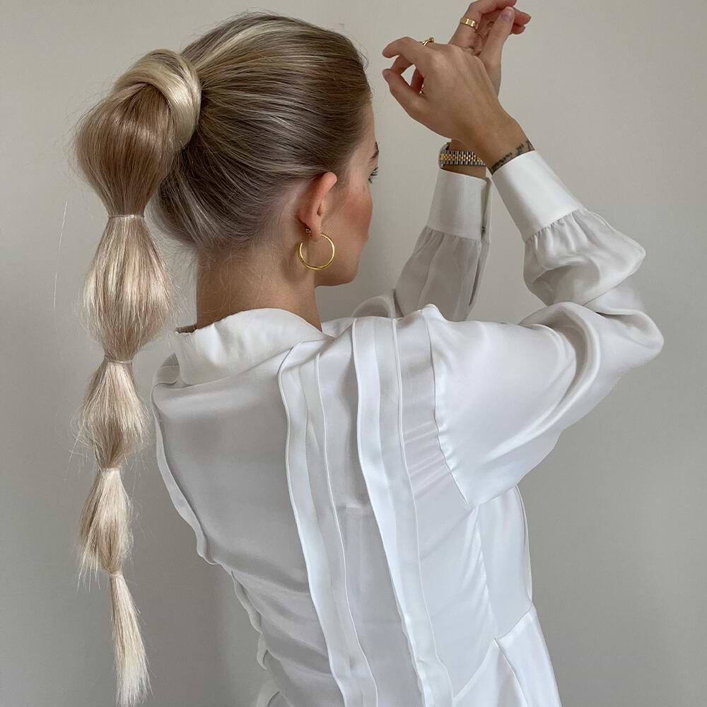 Woman with a long, sectioned ponytail with hair extensions and gold hoop earrings, wearing a white blouse, adjusting her hair against a plain background.