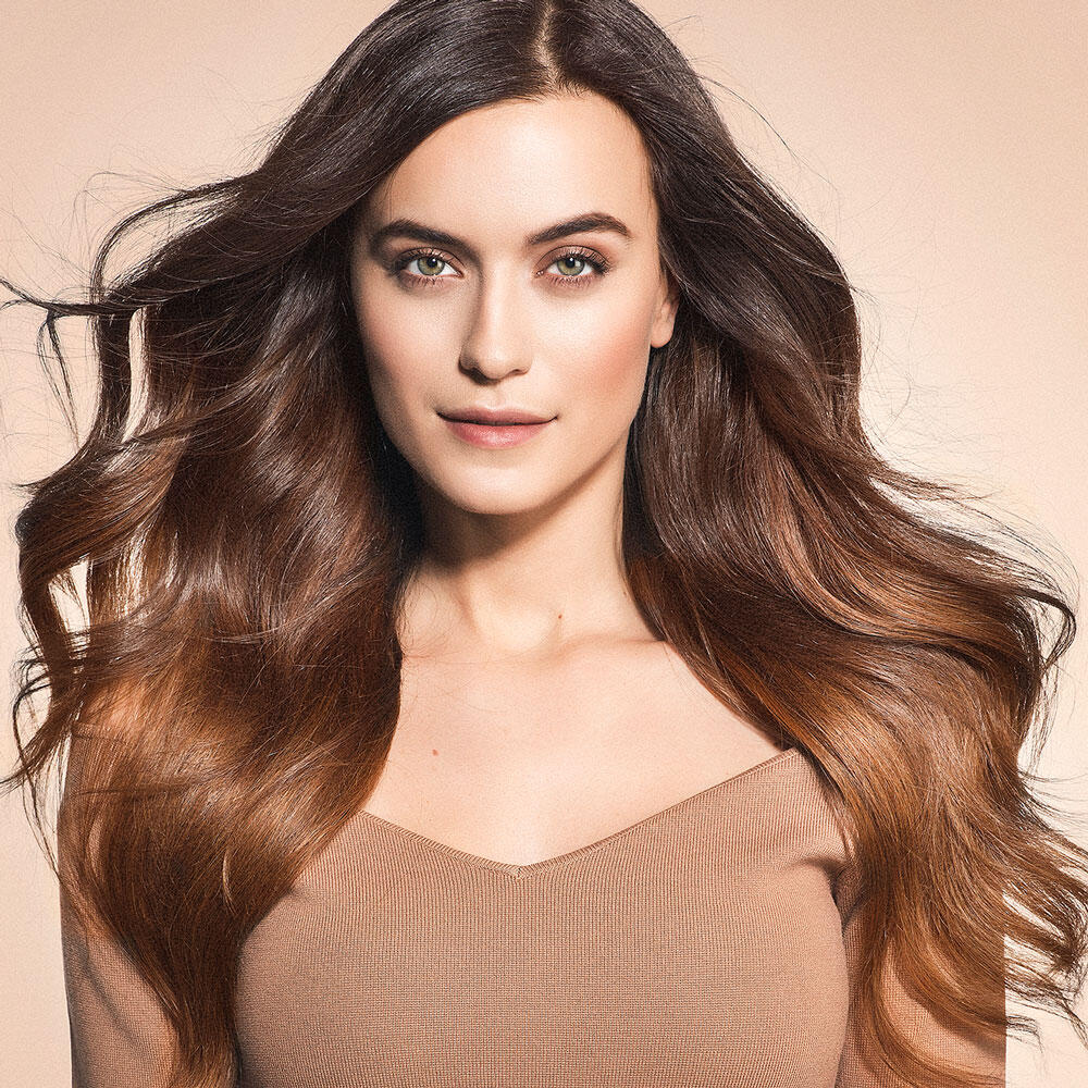 Woman with long, wavy brown hair extensions and a neutral expression, wearing a light brown top, against a beige background.