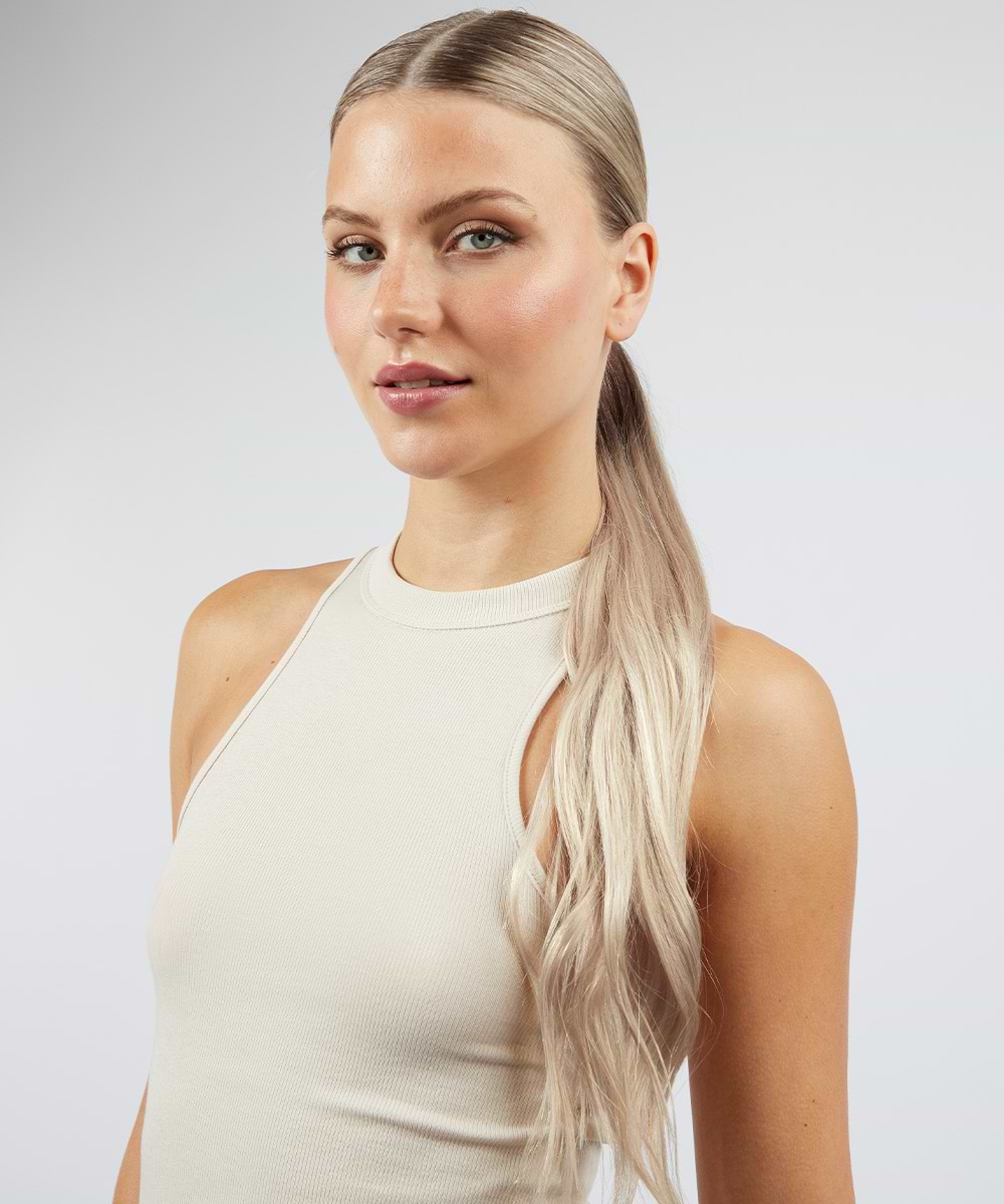 Woman with long, blonde hair extensions in a ponytail, wearing a sleeveless beige top, poses against a plain background.