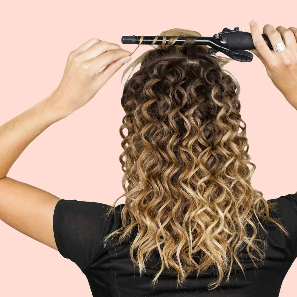 Person using a curling iron to style long, wavy hair extensions against a light pink background.