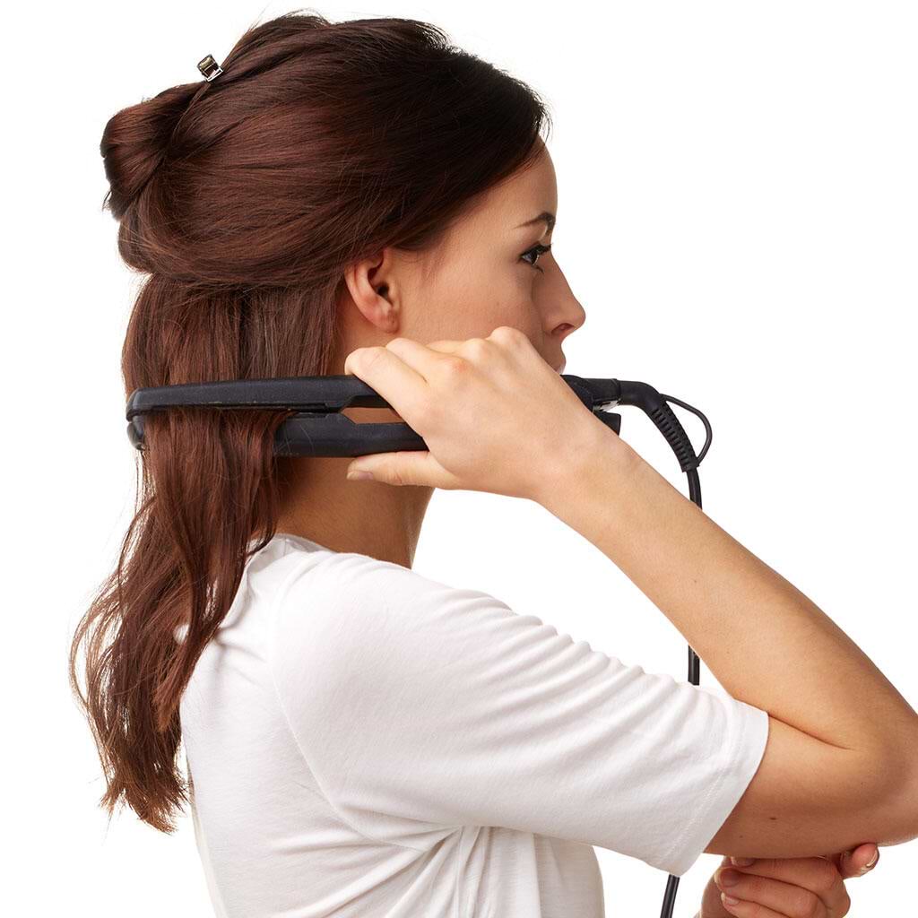 A woman with brown hair extensions straightens her hair using a flat iron. She is wearing a white shirt and is seen from the side.