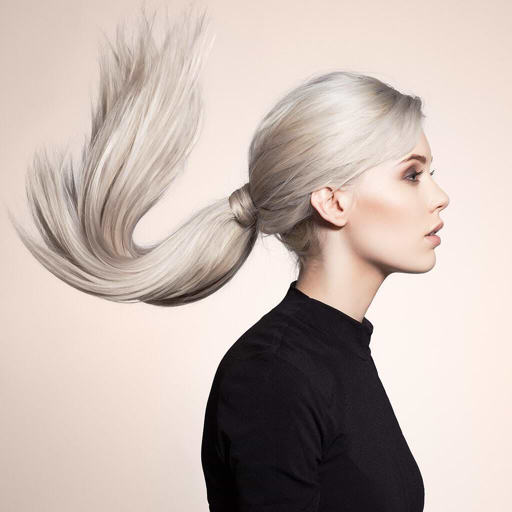 Profile of a person with long, flowing platinum blonde hair extensions in a ponytail, wearing a black top against a neutral background.