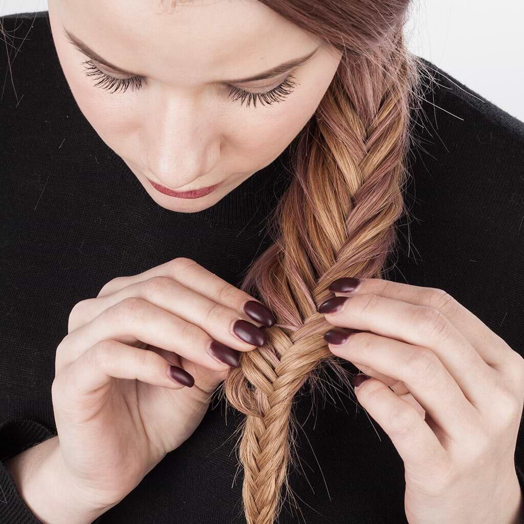 Woman braiding her long, light brown hair extensions into a fishtail braid, wearing a black top, with dark nail polish.