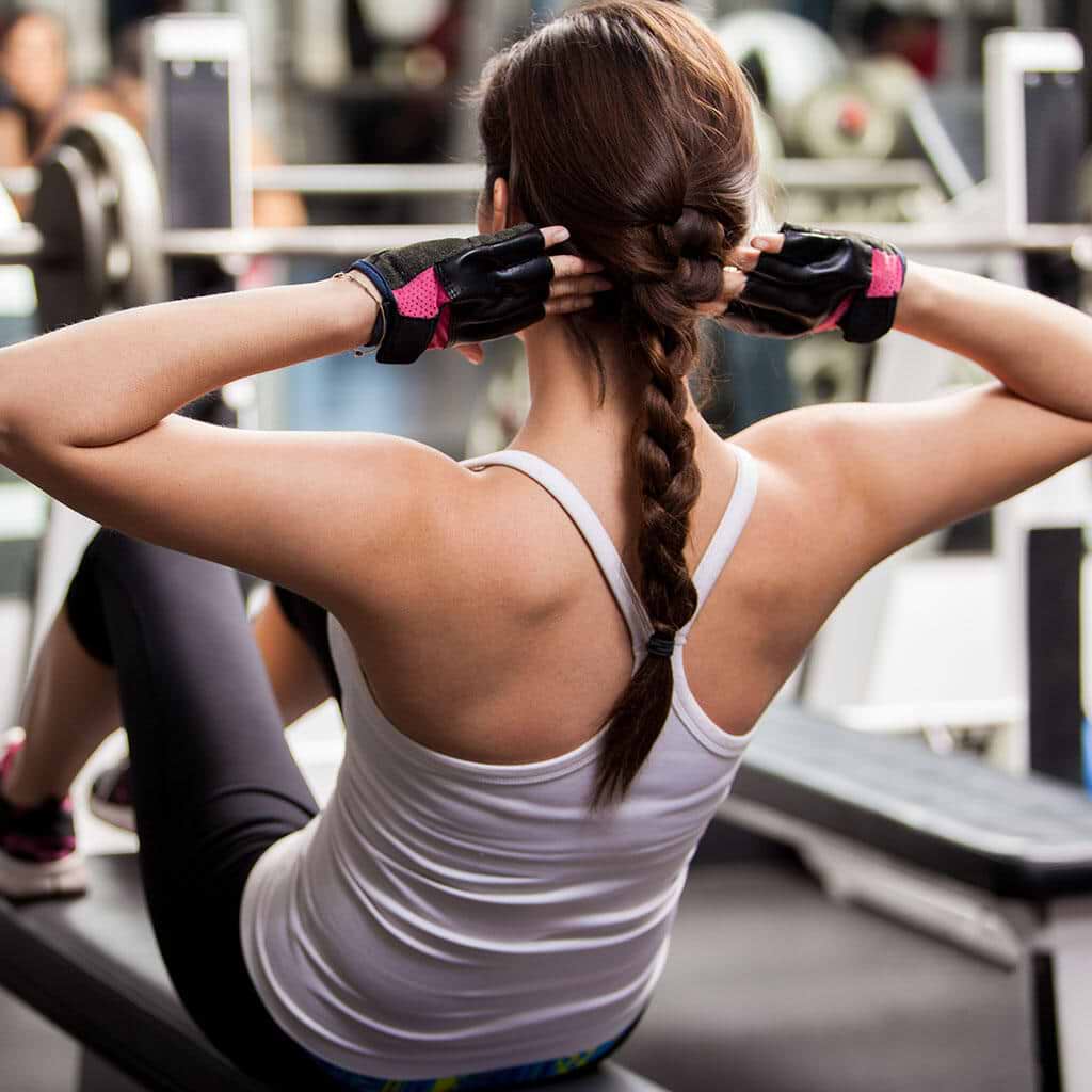 Woman with braided hair extensions doing sit-ups on a bench in a gym, wearing a white tank top and black gloves.
