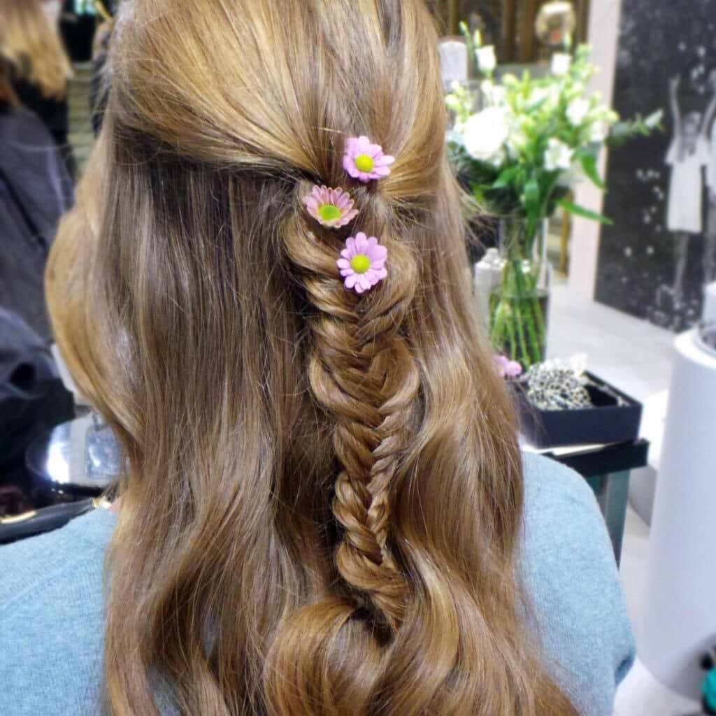 Woman with long, wavy hair extensions styled in a fishtail braid adorned with pink daisy flowers, wearing a blue top in a salon setting.