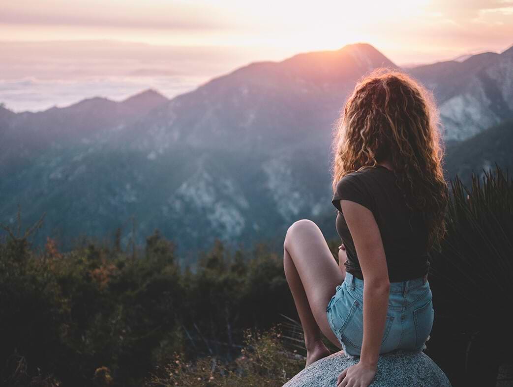A woman with curly hair extensions, sits on a rock, overlooking a mountainous landscape at sunset.
