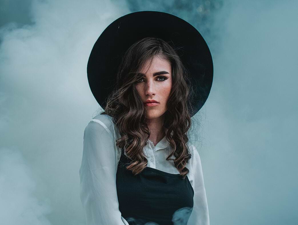 Woman with wavy hair extensions wearing a black hat and white blouse, surrounded by misty smoke.