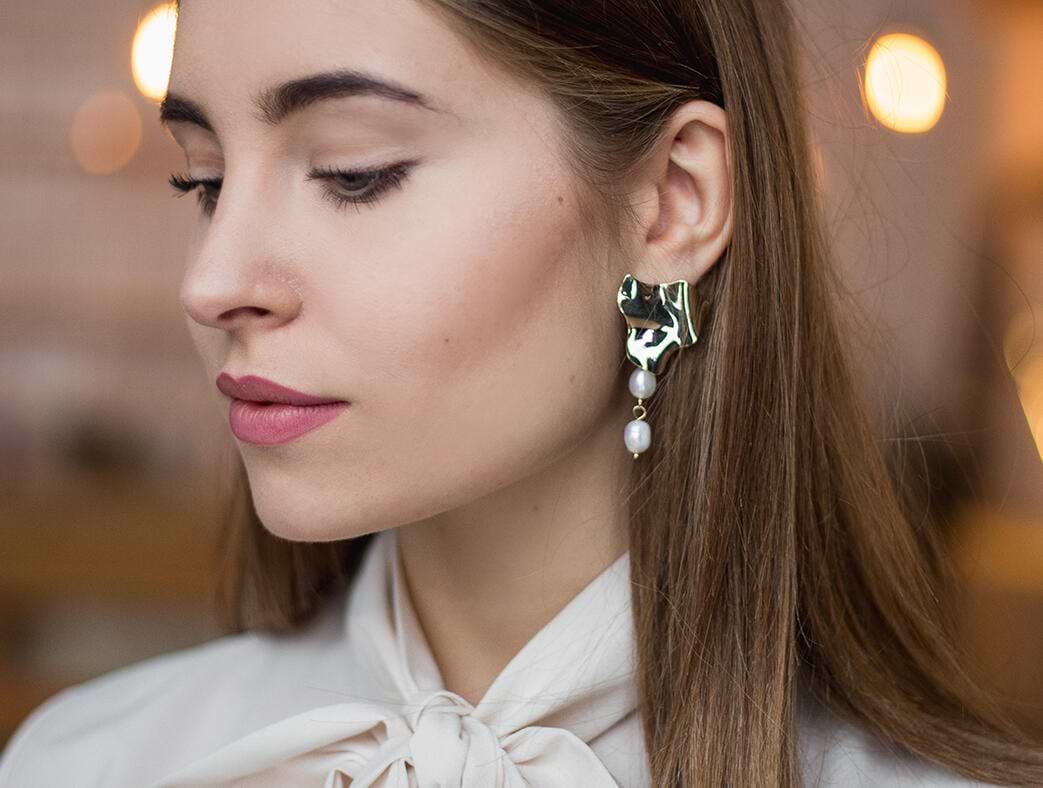 Woman with long brown hair extensions wearing a unique gold and pearl earring, looking down, dressed in a light blouse with a bow tie.