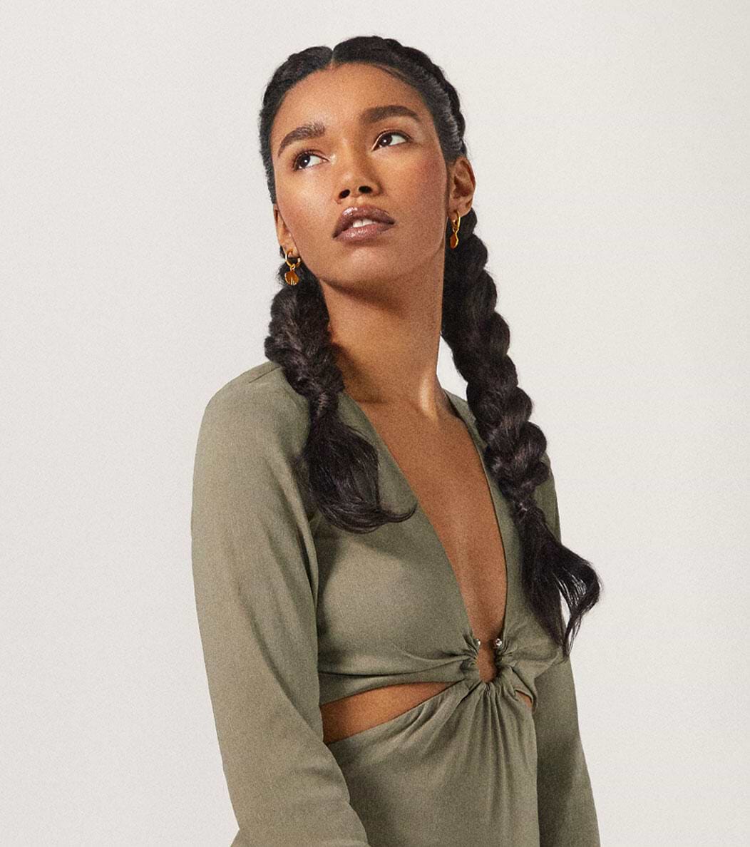 Woman with long braided hair extensions in an olive dress, looking to the side against a plain background.