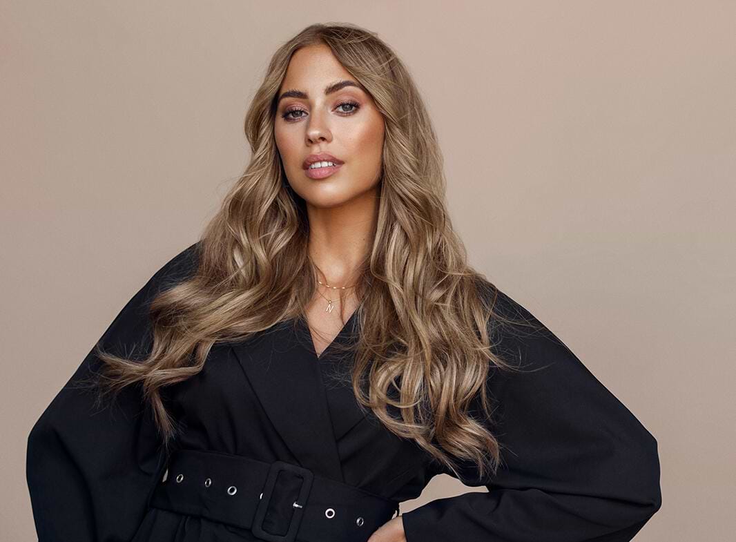 Woman with long, wavy hair extensions wearing a black belted outfit, standing against a beige background.