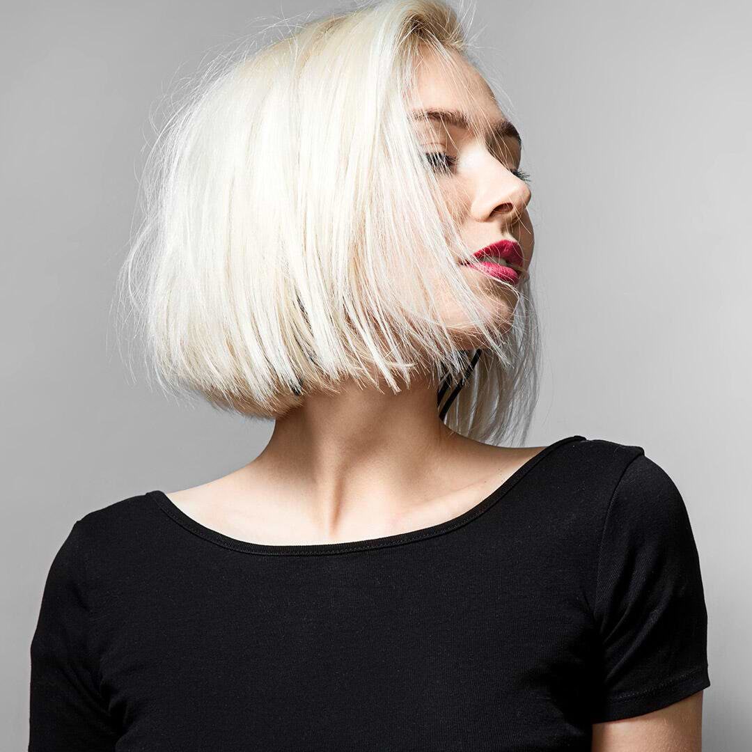 Woman with short blonde bob hairstyle with hair extensions, wearing a black top, looking to the side against a gray background.